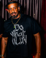 A man wearing a black t-shirt with a white graffiti-style graphic print, styled with a gold chain necklace and headphones, set against a dark curtain background.