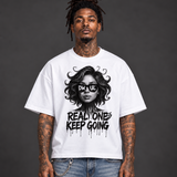 A man with tattoos and dreadlocks stands against a dark gray studio background wearing an oversized white graphic T-shirt with a black illustration of a woman's face and the text 'DW UL REAL ONE KEEP GOING', paired with distressed blue jeans.
