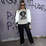 A woman stands in front of a graffiti-covered wall wearing a white zip-up jacket, a beige graphic T-shirt with black text and illustration, black pants, black-and-white sneakers, and sunglasses.