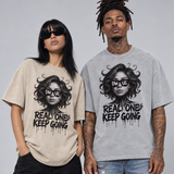Two models wearing matching graphic t-shirts with a black and white illustration of a woman's face and the text 'REAL ONE'S KEEP GOING' printed on the front, posed against a solid gray studio background.