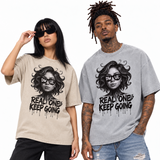 Two models wearing matching graphic t-shirts with a black and white illustration of a woman's face and the text 'REAL ONE KEEP GOING'. The model on the left wears a beige t-shirt with black pants, and the model on the right wears a heather gray t-shirt with distressed blue jeans.