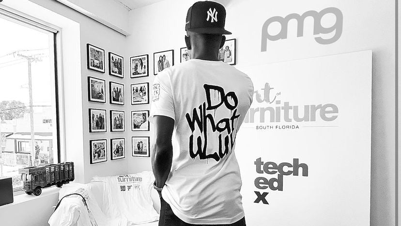 A man stands with his back to the camera in a white room, wearing a white t-shirt with 'Do what u lit' printed on the back and a black New York Yankees baseball cap. The room features a large white display board with 'pmg' and 'tech ed x' branding, and a window on the left.