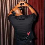 A man seen from behind wearing a black t-shirt with a small red heart graphic on the back, posing with his hands forming a heart shape over his head in front of brown and red curtains.