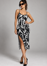 A woman modeling a black spaghetti-strap midi dress with a bold white graffiti-style abstract print, styled with black sunglasses and black high heels against a neutral gray studio background.
