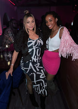 Two women posing together in a dimly lit nightclub or bar. The woman on the left wears a black faux-fur shrug over a black and white graphic dress, and the woman on the right wears a white crop top, a pink fringe shrug, and a hot pink mini skirt.